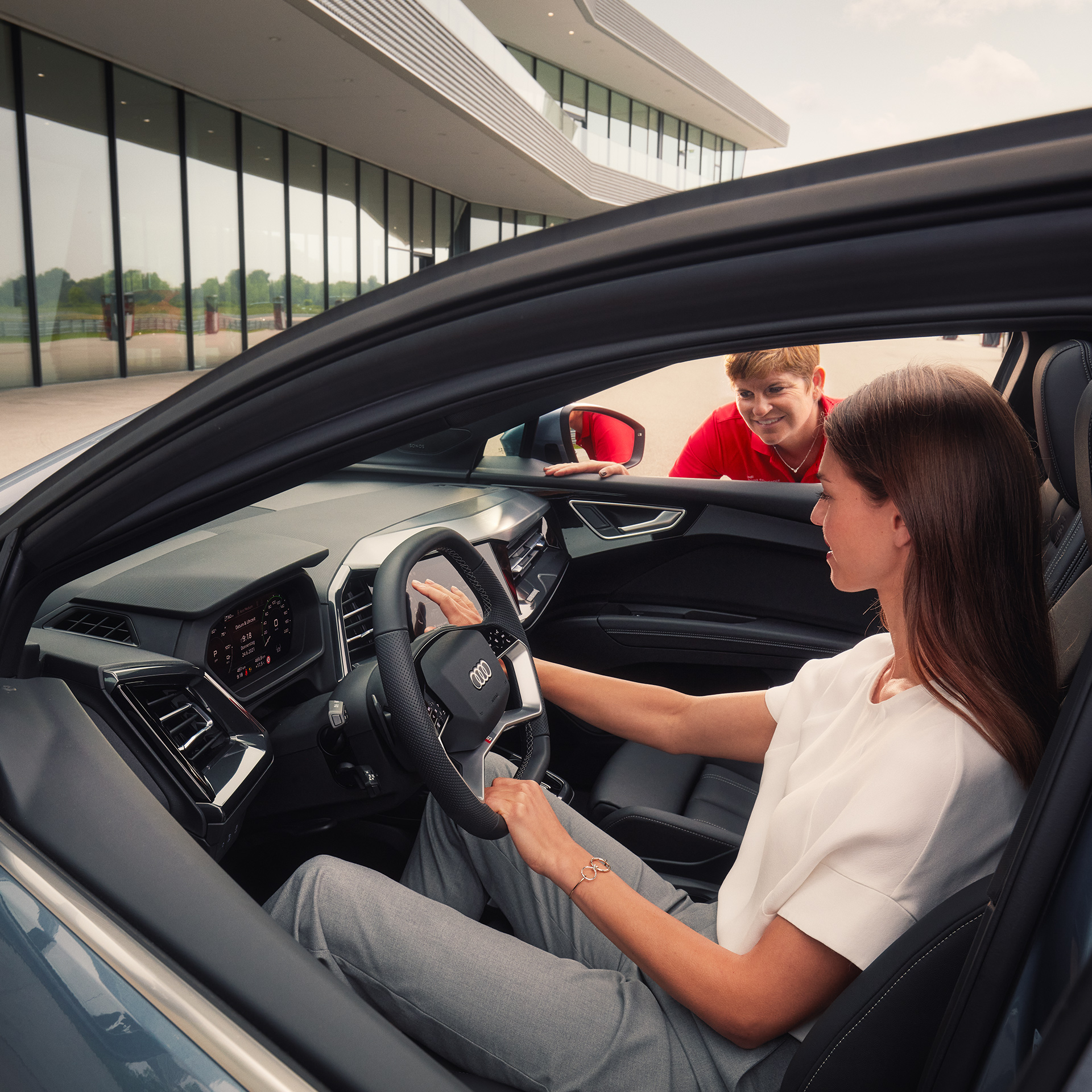 Woman with dark long hair sits in an Audi model, on the passenger side a woman in red t-shirt looks through the window and explains the vehicle