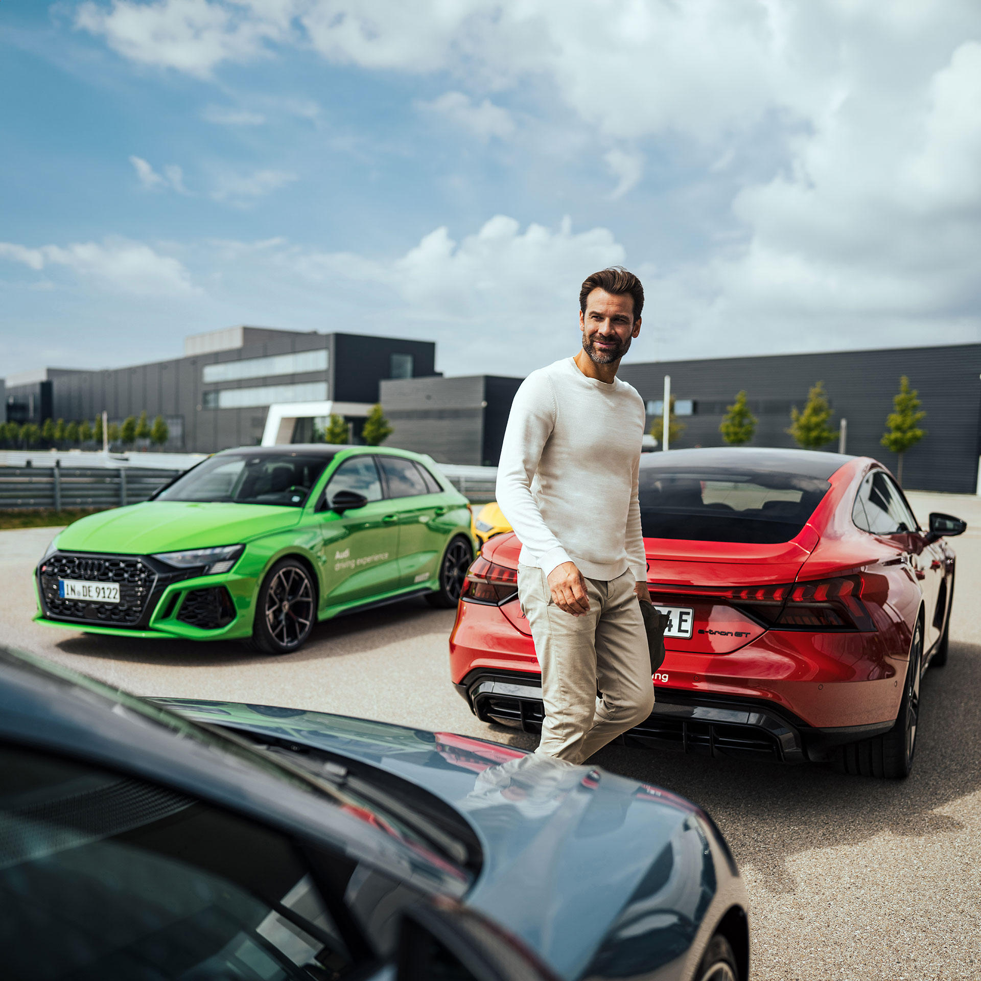 A man walks between three cars, on the left is a light green Audi model, to the right is a red Audi RS e-tron GT and in front is another dark grey Audi model.